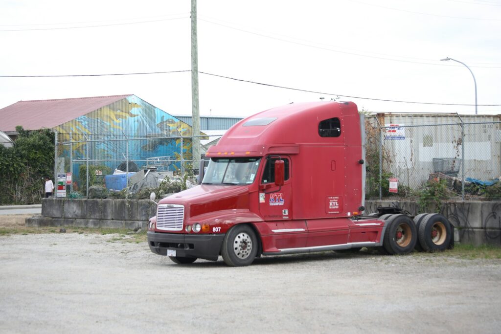 A red semi truck parked on a gravel lot near a chain link fence
