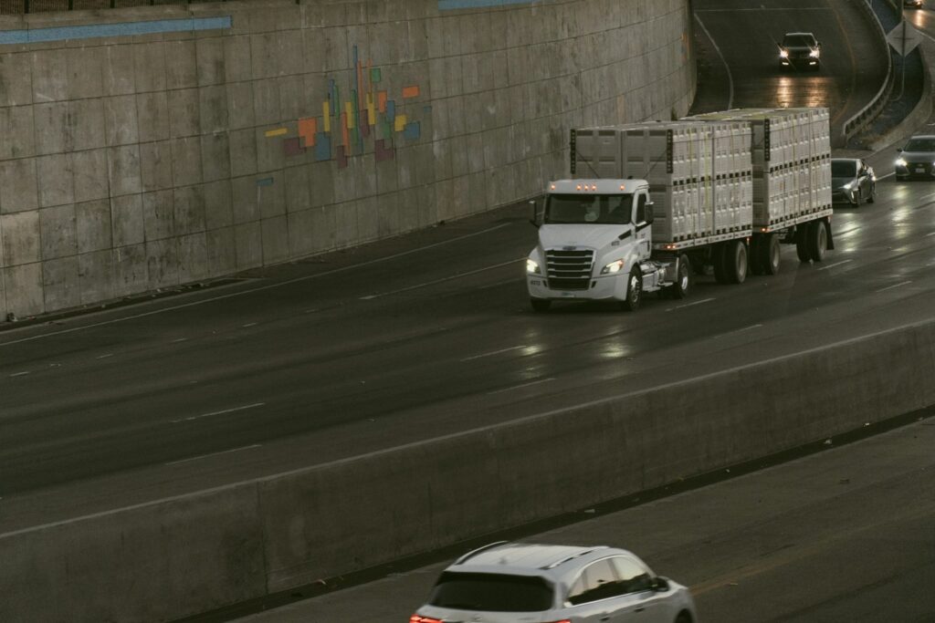 A semi-truck carrying white cargo containers on a highway beside a concrete wall