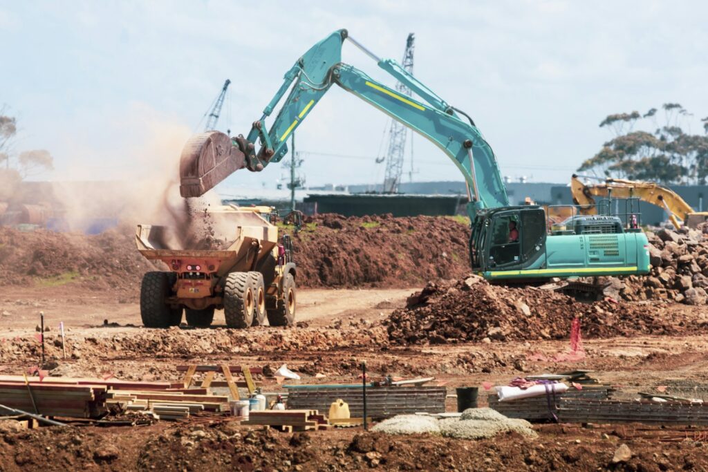 A teal excavator dumping soil into a yellow dump truck at a construction site